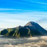 Snow-capped mountain peak rising above a blanket of clouds with a bright blue sky above.