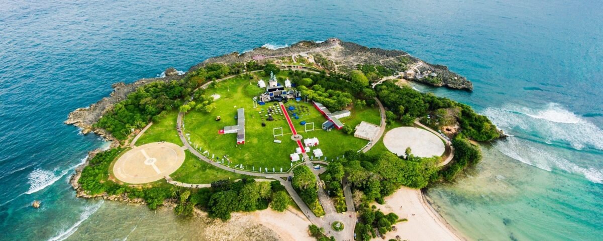 Aerial view of a small island park with a circular green lawn, paths, and event tents surrounded by blue ocean and rocky shores.