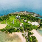 Aerial view of a small island park with a circular green lawn, paths, and event tents surrounded by blue ocean and rocky shores.