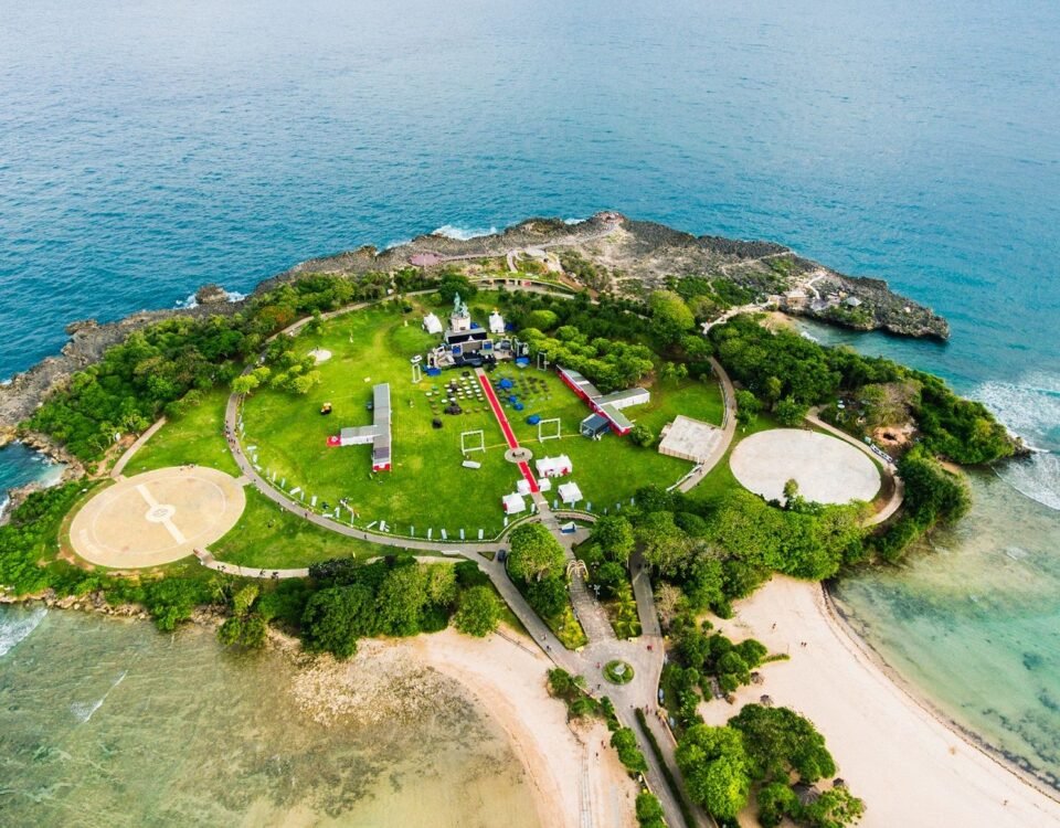 Aerial view of a small island park with a circular green lawn, paths, and event tents surrounded by blue ocean and rocky shores.