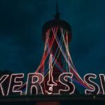 Nighttime scene with a tall tower and a neon sign across a rooftop; multicolor light trails descend from the tower and outline letters across the sign.
