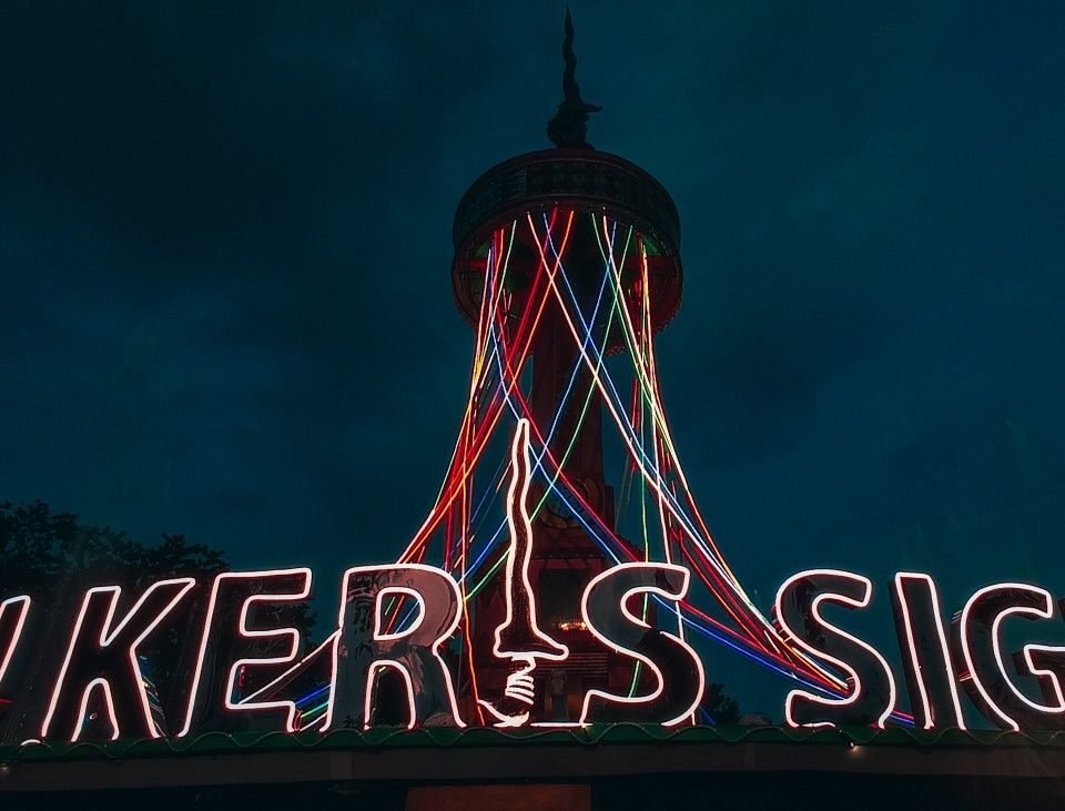 Nighttime scene with a tall tower and a neon sign across a rooftop; multicolor light trails descend from the tower and outline letters across the sign.