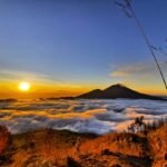 Sunrise over a sea of clouds with a silhouetted volcano on the horizon and golden grasses in the foreground; warm colors dominate the scene.
