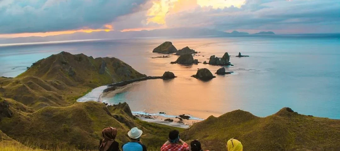 Five people sit on a grassy hill, watching a sunset over a rocky coastline and small islets in calm turquoise water distance view.
