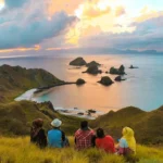 Five people sit on a grassy hill, watching a sunset over a rocky coastline and small islets in calm turquoise water distance view.