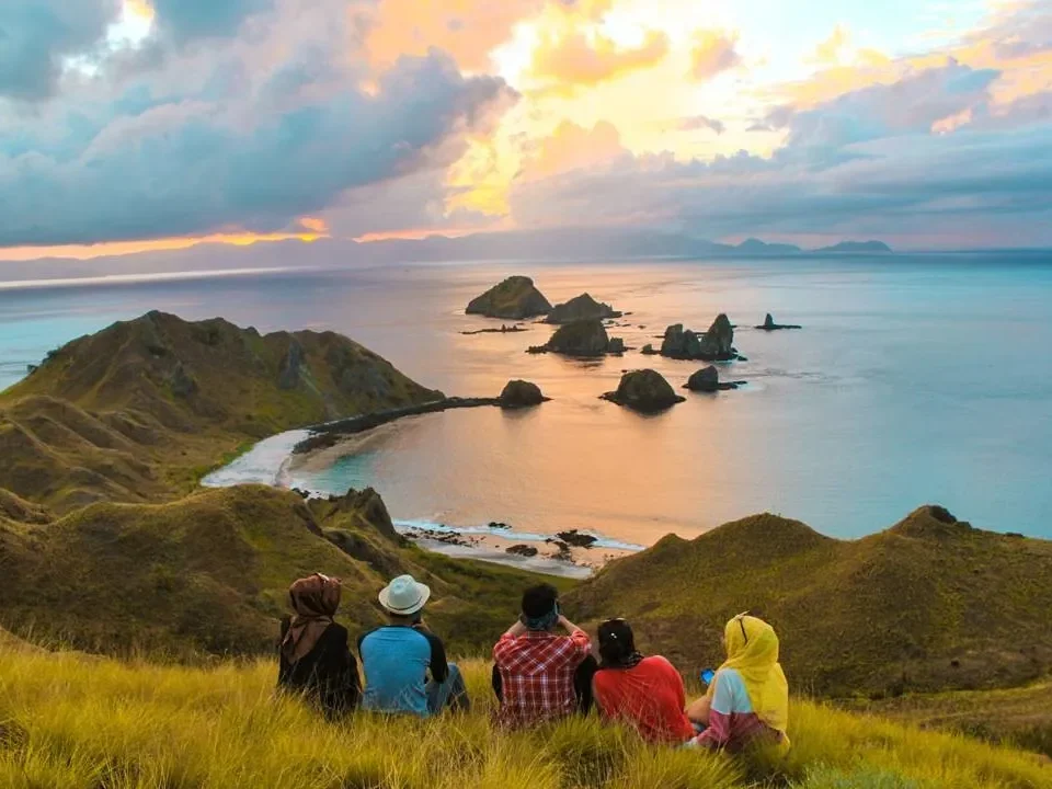 Five people sit on a grassy hill, watching a sunset over a rocky coastline and small islets in calm turquoise water distance view.