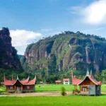 Rural landscape with green fields, traditional curved-roof wooden houses, and towering limestone cliffs under a blue sky.