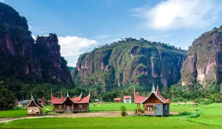 Rural landscape with green fields, traditional curved-roof wooden houses, and towering limestone cliffs under a blue sky.
