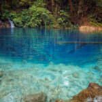 Clear tropical lake with turquoise water and rocky shoreline, dense green jungle along the banks and a small waterfall on the left.