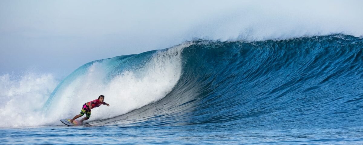 Surfer in a red shirt and colorful shorts rides a large blue curling wave.