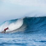 Surfer in a red shirt and colorful shorts rides a large blue curling wave.
