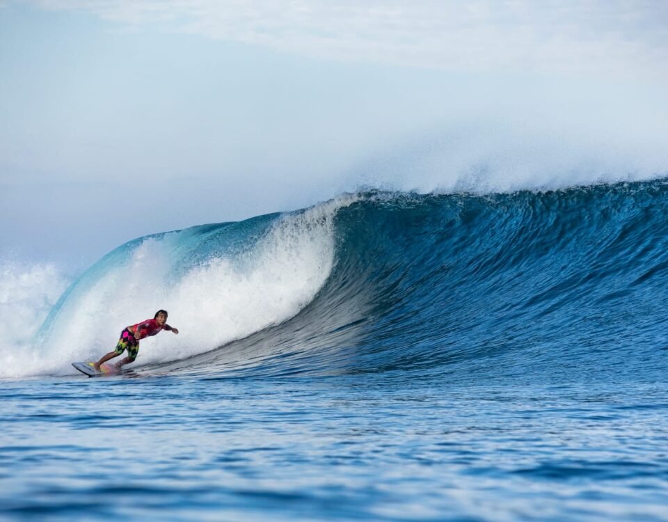 Surfer in a red shirt and colorful shorts rides a large blue curling wave.
