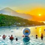 Sunset Point Bali infinity pool with swimmers, overlooking the sea at sunset and a distant volcanic hill.