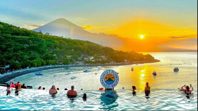 Sunset Point Bali infinity pool with swimmers, overlooking the sea at sunset and a distant volcanic hill.