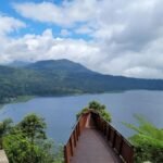Wooden viewing walkway extends toward a calm lake framed by green hills under a partly cloudy sky.
