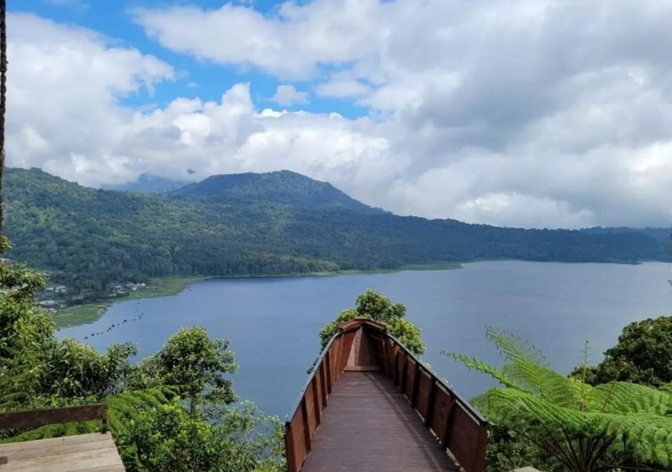 Wooden viewing walkway extends toward a calm lake framed by green hills under a partly cloudy sky.