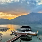 Sunset over a calm lake with a mountain range in the distance and boats docked along wooden piers.