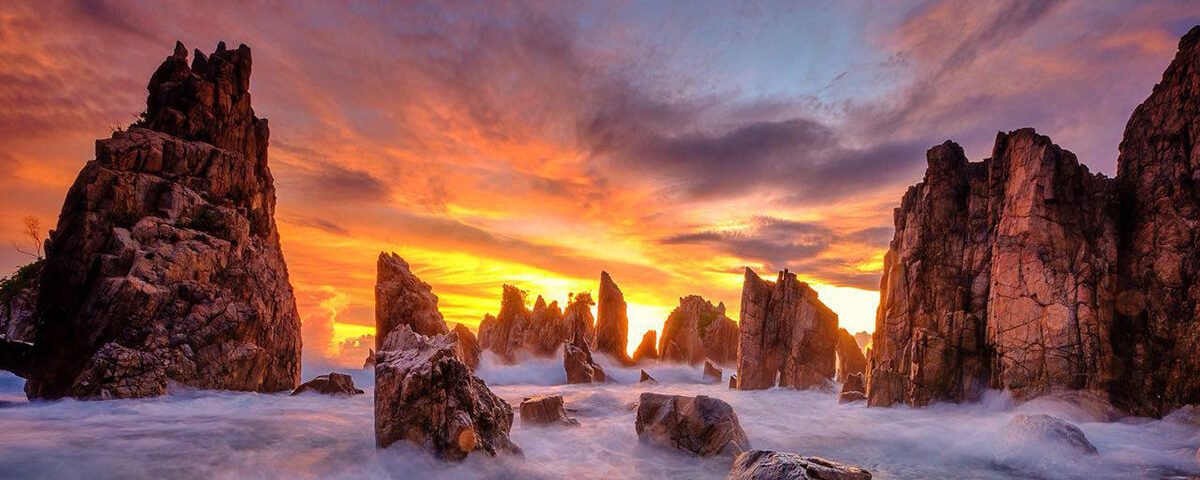 Rugged sea stacks rise from foaming waters at sunset, with a fiery orange sky and purple clouds above the horizon.