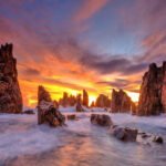 Rugged sea stacks rise from foaming waters at sunset, with a fiery orange sky and purple clouds above the horizon.