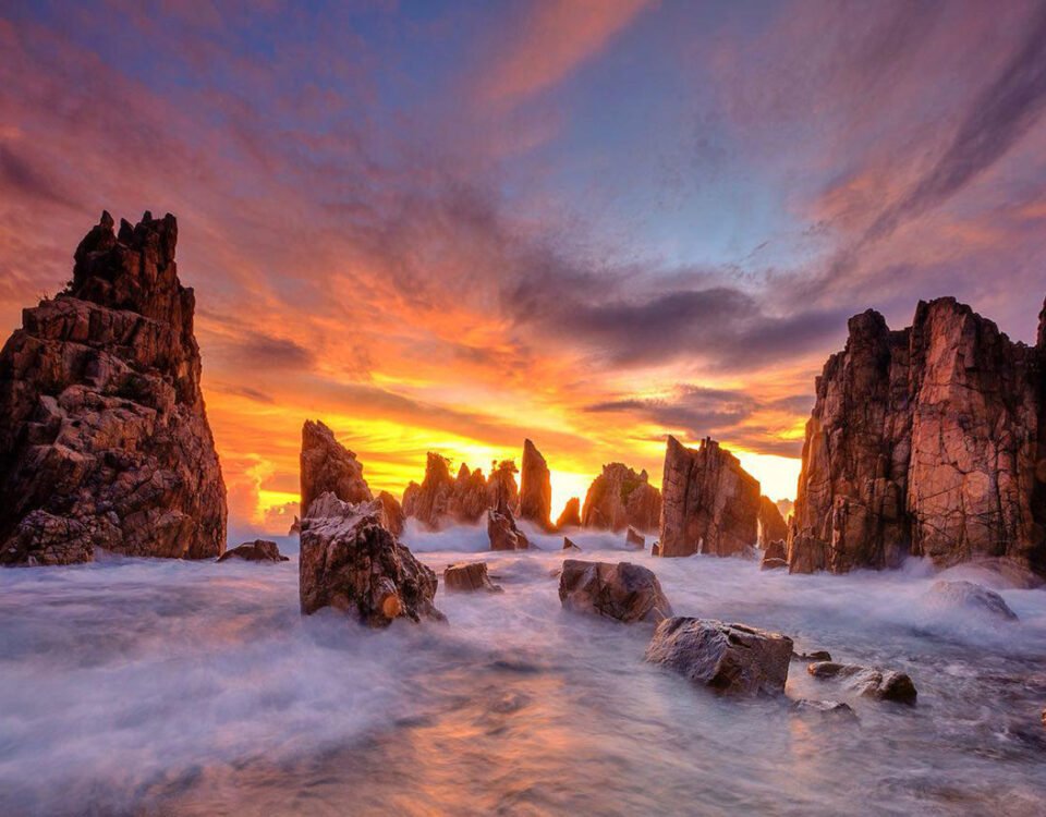 Rugged sea stacks rise from foaming waters at sunset, with a fiery orange sky and purple clouds above the horizon.