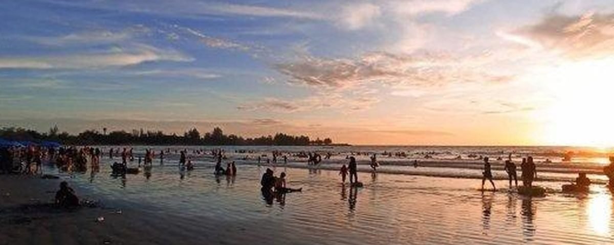 Crowded beach at sunset with people wading and playing in shallow surf while the sky glows orange and blue in the background.