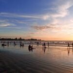 Crowded beach at sunset with people wading and playing in shallow surf while the sky glows orange and blue in the background.