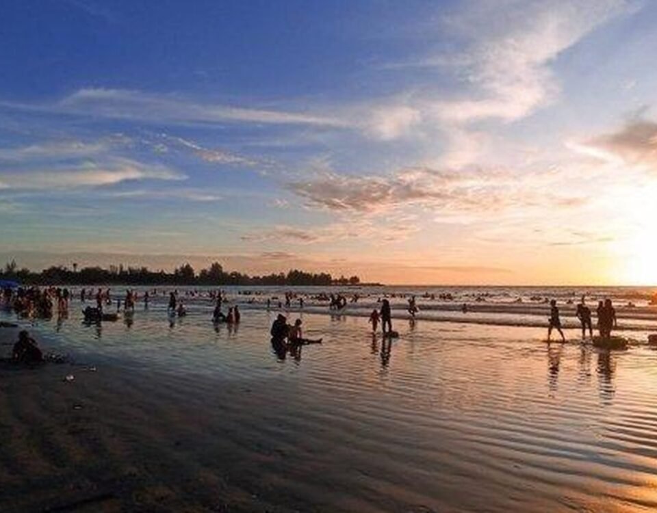 Crowded beach at sunset with people wading and playing in shallow surf while the sky glows orange and blue in the background.
