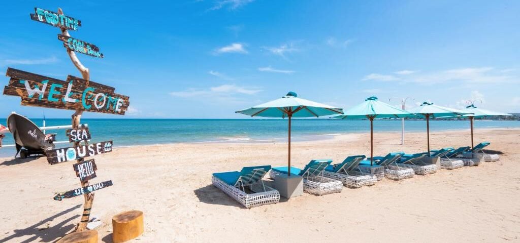 Beach scene with a weathered sign and a row of blue lounge chairs under turquoise umbrellas on sandy shore by calm blue water on a sunny day.