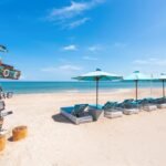 Beach scene with a weathered sign and a row of blue lounge chairs under turquoise umbrellas on sandy shore by calm blue water on a sunny day.