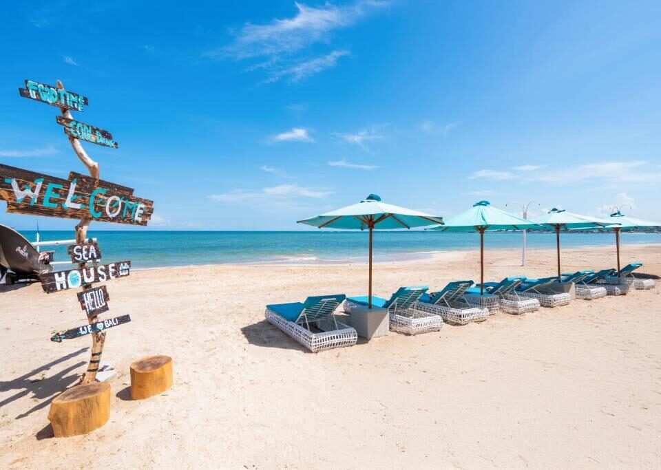 Beach scene with a weathered sign and a row of blue lounge chairs under turquoise umbrellas on sandy shore by calm blue water on a sunny day.