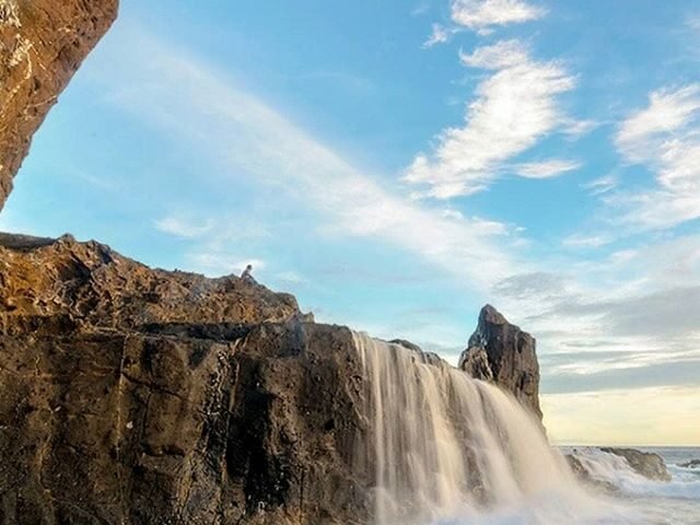 Rocky coastline waterfall pouring over cliffs into blue-green sea; a person stands on the edge watching the water fall.