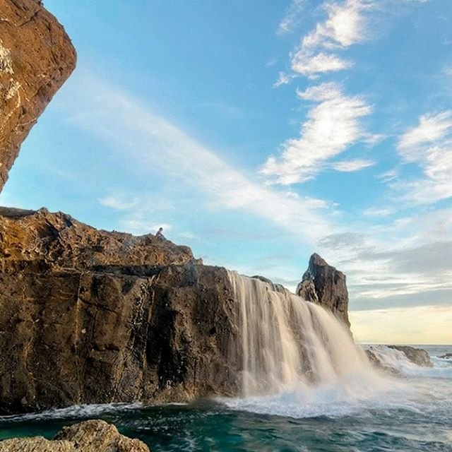 Rocky coastline waterfall pouring over cliffs into blue-green sea; a person stands on the edge watching the water fall.
