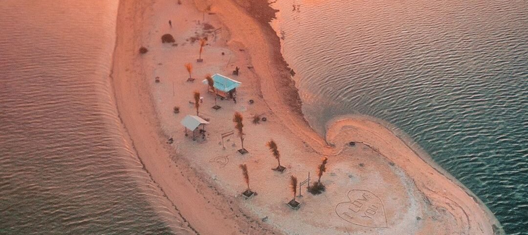 Crescent-shaped sandy island at sunset with palm trees, beach umbrellas and a heart carved in the sand, and a boat nearby in calm turquoise water.