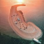 Crescent-shaped sandy island at sunset with palm trees, beach umbrellas and a heart carved in the sand, and a boat nearby in calm turquoise water.