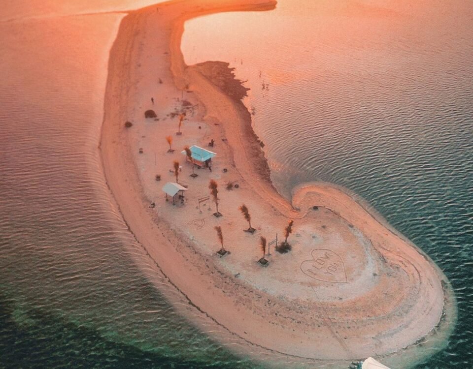 Crescent-shaped sandy island at sunset with palm trees, beach umbrellas and a heart carved in the sand, and a boat nearby in calm turquoise water.