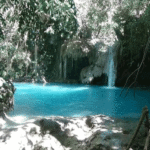 Turquoise natural pool with a small waterfall surrounded by dense jungle and rocks.