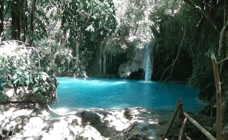 Turquoise natural pool with a small waterfall surrounded by dense jungle and rocks.