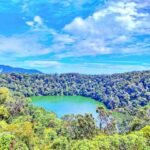 Bright blue sky over a dense green forest surrounding a small turquoise lake at center.