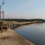 Wooden pier extends into calm water beside a small hut and benches on a concrete quay cresting with a signpost and streetlight.
