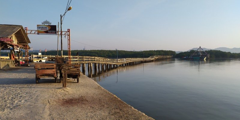 Wooden pier extends into calm water beside a small hut and benches on a concrete quay cresting with a signpost and streetlight.