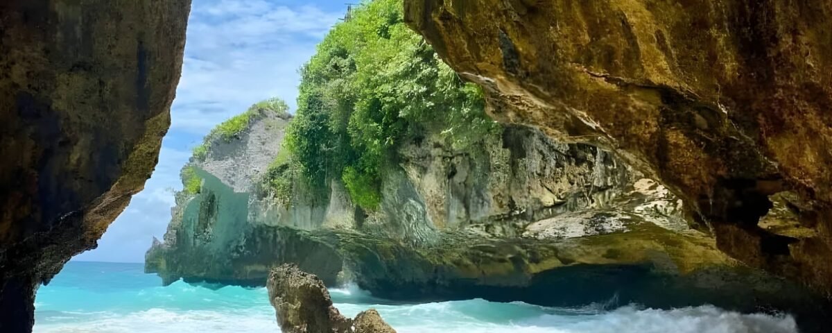 A sea cave opening to turquoise water, with sandy shore and rugged rock walls covered in green vegetation on a sunny day.