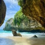 A sea cave opening to turquoise water, with sandy shore and rugged rock walls covered in green vegetation on a sunny day.