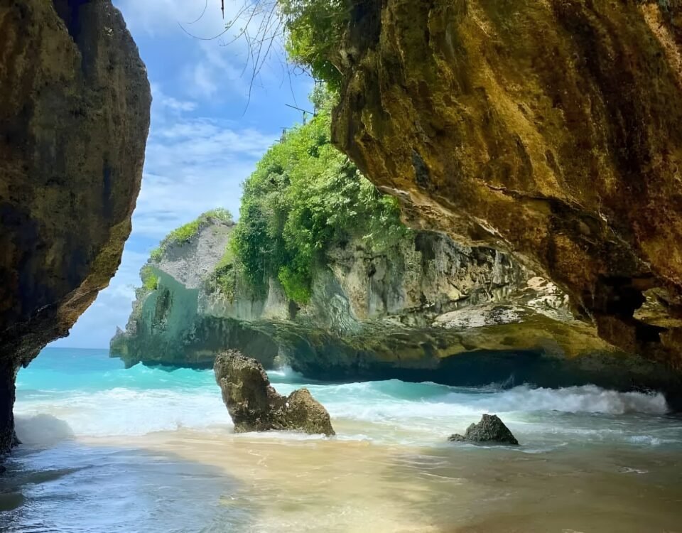 A sea cave opening to turquoise water, with sandy shore and rugged rock walls covered in green vegetation on a sunny day.