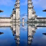 Stone gate with ornate carvings split by a narrow gap, a person standing between the halves with arms raised, reflected in a calm pool with a distant mountain and blue sky behind.