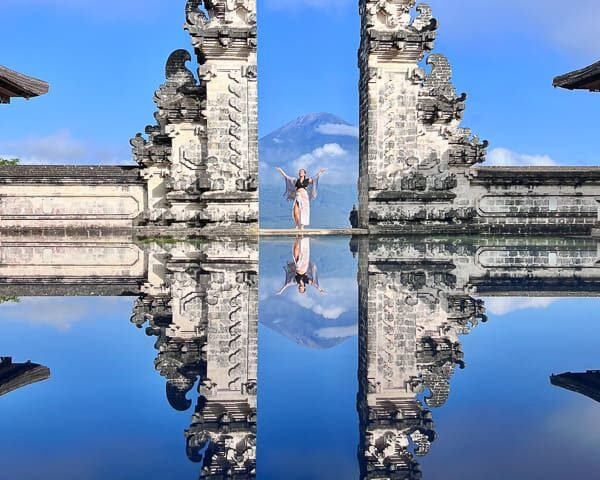 Stone gate with ornate carvings split by a narrow gap, a person standing between the halves with arms raised, reflected in a calm pool with a distant mountain and blue sky behind.