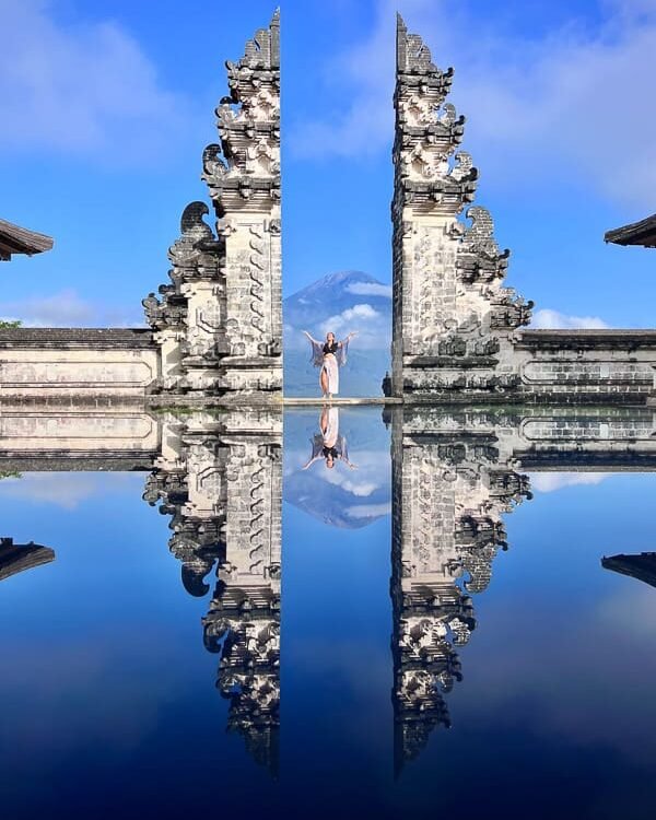 Stone gate with ornate carvings split by a narrow gap, a person standing between the halves with arms raised, reflected in a calm pool with a distant mountain and blue sky behind.