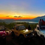 Sunset over a valley and lake with distant mountains, a row of parked off-road jeeps in the foreground and people viewing the scene (silhouettes).