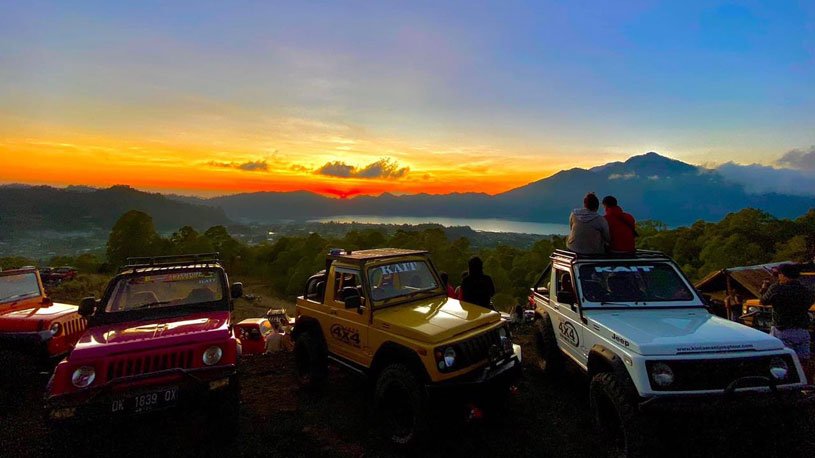 Sunset over a valley and lake with distant mountains, a row of parked off-road jeeps in the foreground and people viewing the scene (silhouettes).