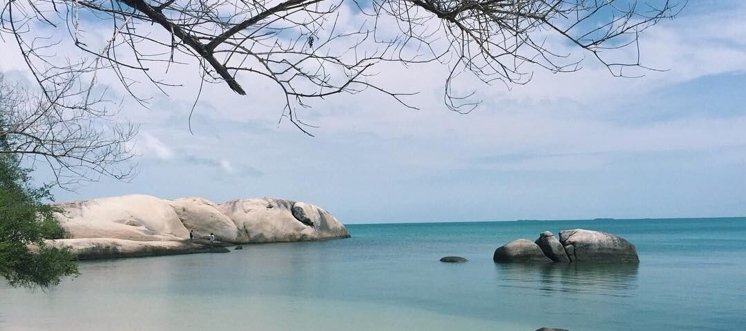 Peaceful tropical beach with pale sand, calm turquoise water, and large granite rocks along the shoreline under a blue sky.