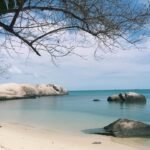 Peaceful tropical beach with pale sand, calm turquoise water, and large granite rocks along the shoreline under a blue sky.